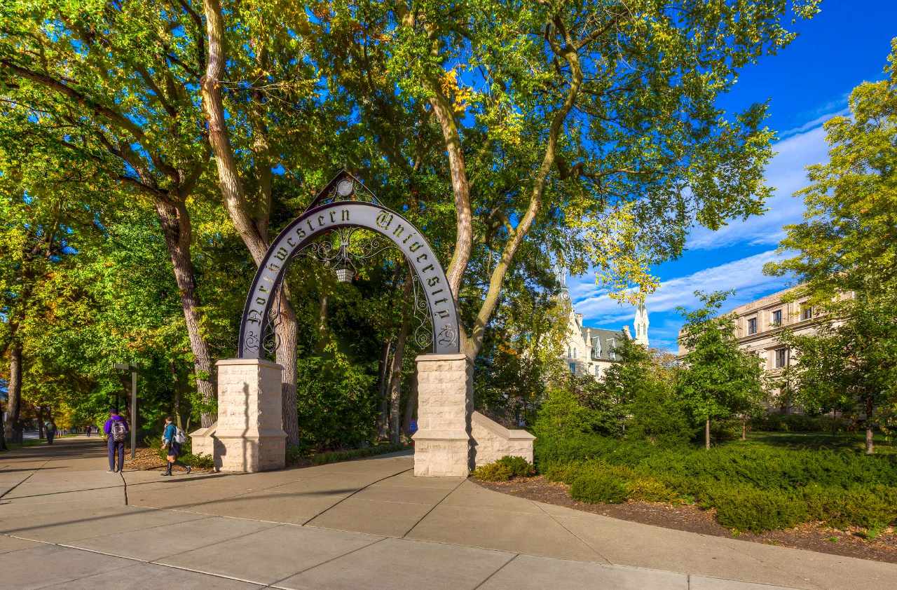 The Northwestern arch at the corner of Sheridan Rd and Chicago Ave with trees and bushes behind it and a couple of students walking past