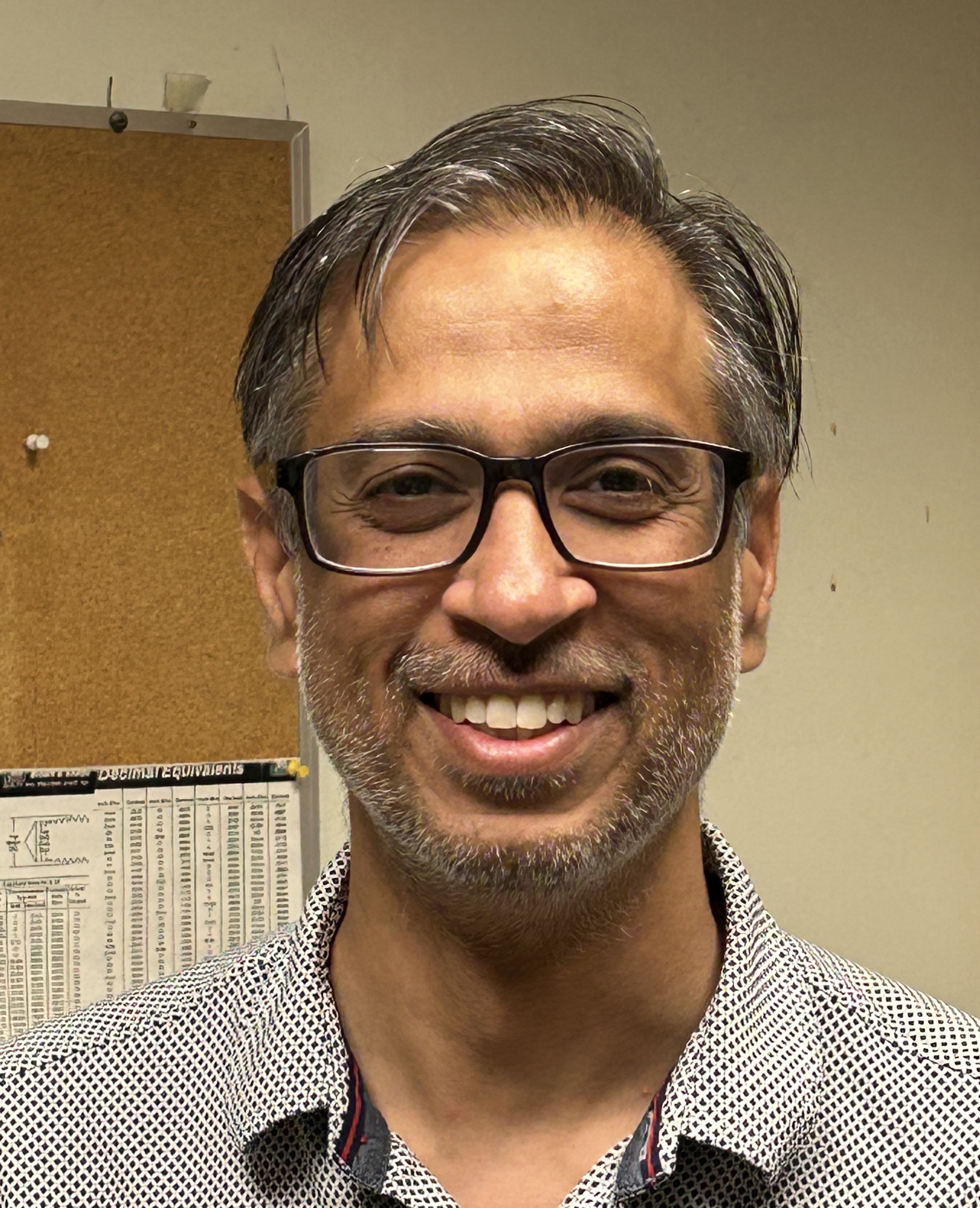 Headshot of Sumit smiling with a corkboard and tan wall in the background